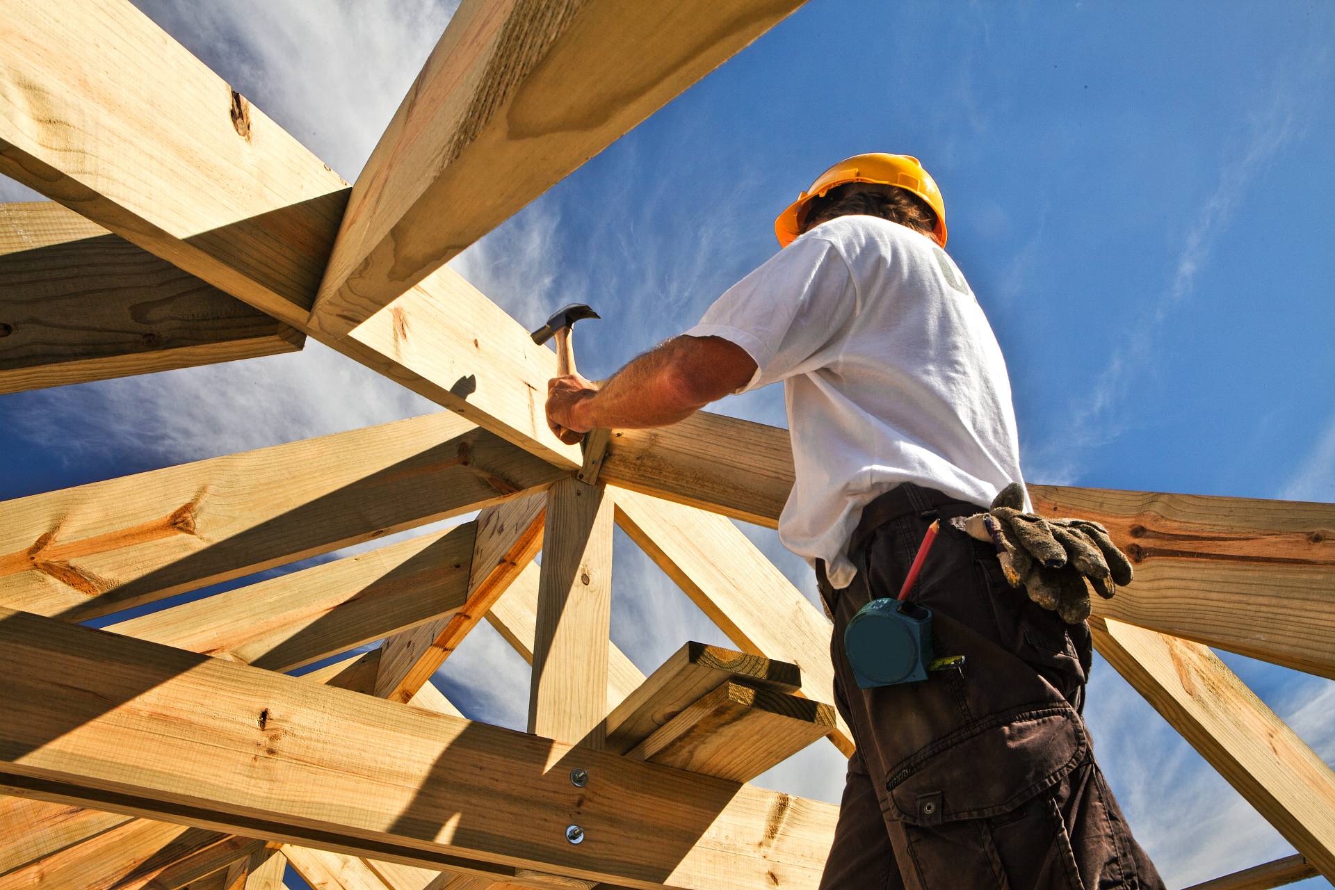Handwerker mit Schutzhelm schlägt einen Nagel in die Holzbalken eines Dachstuhls, mit Blick in den Himmel.