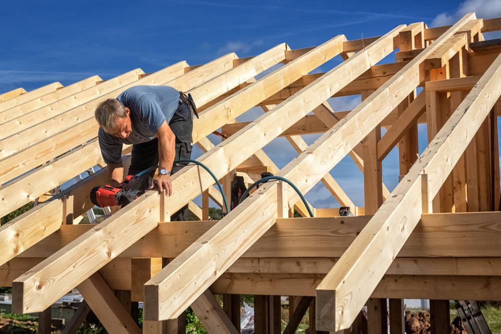 Zimmermann arbeitet mit einer Druckluftpistole an der Dachkonstruktion eines Holzhauses unter blauem Himmel.