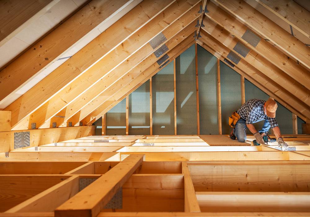 Arbeiter verlegt Dämmplatten im Spitzboden eines neu errichteten Holzhauses mit sichtbarer Dachkonstruktion.